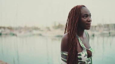 Afrika örgülü güzel bir kadın limanda iskele boyunca yürüyor ve deniz manzarasına bakıyor.