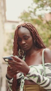 Sokakta üzerinde Afrika örgüsü olan güzel bir kadın ayakta dikiliyor ve cep telefonu kullanıyor. Şık kız akıllı telefondan mesaj yazıyor.
