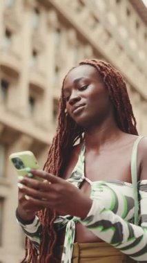 Afrika örgülü, sırıtan muhteşem bir kadın kaldırımın kenarındaki kaldırımda duruyor ve cep telefonu kullanıyor. Şık kız akıllı telefondan mesaj yazıyor.