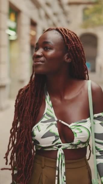 垂直ビデオ 上の通りを歩くに身を包んだアフリカの編組を持つ豪華な女性 スタイリッシュな笑顔の女の子は 建物の上階を見て — ストック動画