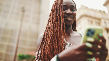 Üzerinde Afrika örgüleri olan gülümseyen muhteşem bir kadın sokakta dikiliyor ve cep telefonu kullanıyor. Şık kız akıllı telefondan mesaj yazıyor.
