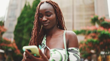 Üzerinde Afrika örgüleri olan gülümseyen muhteşem bir kadın sokakta dikiliyor ve cep telefonu kullanıyor. Şık kız akıllı telefondan mesaj yazıyor.