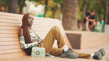 Profile göre, Afrika örgülü muhteşem bir kadın üst tarafta oturuyor ve pantolonu da bankta dinleniyor.