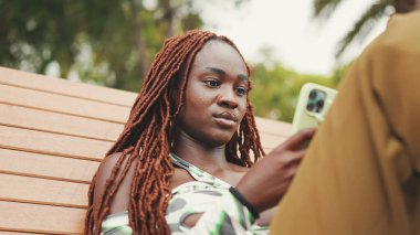 Üzerinde Afrika örgüleri olan gülümseyen muhteşem kadın bankta otururken cep telefonu kullanıyor. Şık bir kız fotoğraf izliyor, akıllı telefonunda videosu var.