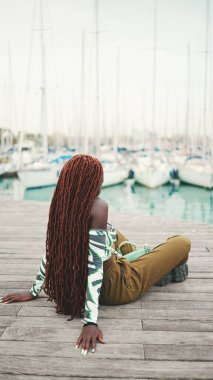 Üstü örülü Afrikalı bir kadın limandaki yat ve gemilere bakıyor. Arka plan