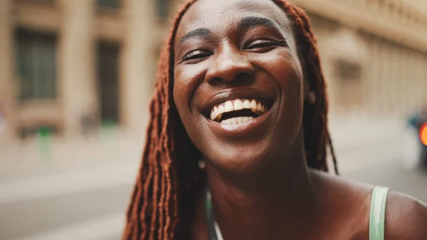 Close-up of beautiful woman with African braids raising her head and looking at the camera with smile on the building background.