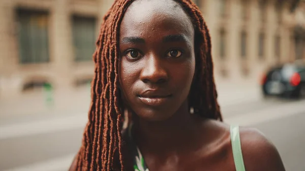 Close-up of beautiful woman with African braids raising her head and looking at the camera with smile on the building background.