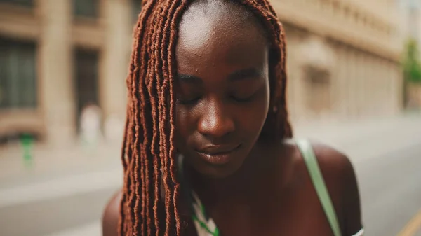 Close-up of beautiful woman with African braids raising her head and looking at the camera with smile on the building background.