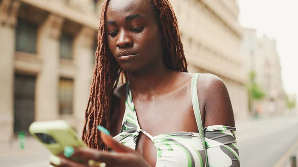 Smiling gorgeous woman with African braids wearing top stands on the sidewalk next to the road and uses mobile phone. Stylish girl writes message on smartphone