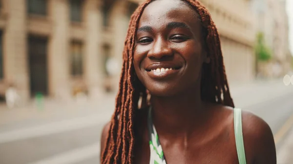 Close-up of beautiful woman with African braids raising her head and looking at the camera with smile on the building background.