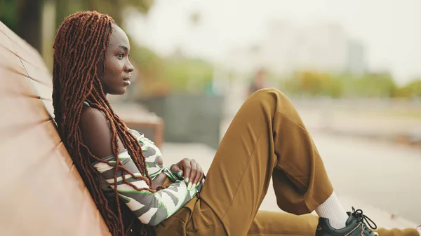 Profile gorgeous woman with African braids wearing in top and trousers is resting sitting on bench