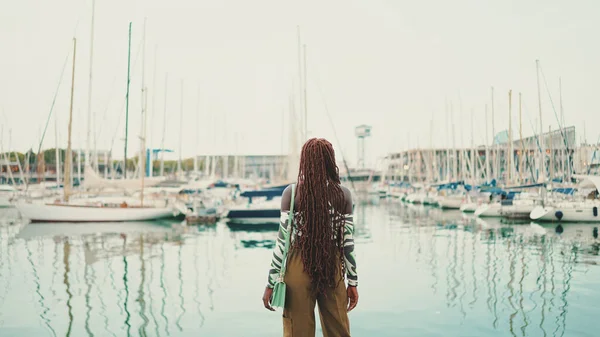 港の桟橋に立っているヨットや船を見て上を着てアフリカの編組を持つ女性 — ストック写真