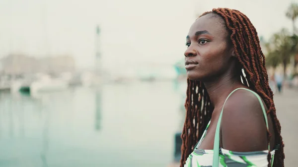 Woman with African braids wearing top looks at the yachts and ships standing on the pier in the port.