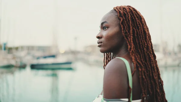 Woman with African braids wearing top looks at the yachts and ships standing on the pier in the port.