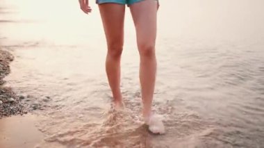 Sahilde çıplak ayakla yürüyen ince bacaklı genç bir kadın. İnce kadın bacakları ve ayakları kumlu sahildeki deniz suyu dalgaları boyunca yürüyor.