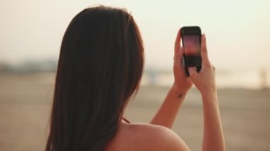 Uzun saçlı güzel kahverengi saçlı kadın sahilde dururken cep telefonu kullanıyor. Güzel kız akıllı telefondan fotoğraf çekiyor. Ağır çekim. Arka plan