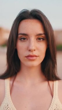 Yakın plan, kumsalda oturan kahverengi saçlı, uzun saçlı güzel bir kadın var. Çilli ve uzun kirpikli güzel bir kız kameraya bakıyor ve gülümsüyor. yavaş çekim