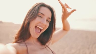 Uzun saçlı, güzel, kahverengi saçlı kadın cep telefonu kullanıyor. Mayo giyen gülümseyen kız deniz manzaralı sahilde selfie çekiyor. Arkaplan