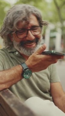 VERTICAL VIDEO: Smiling middle-aged man with gray hair and beard sits on bench and uses mobile phone. Mature man in eyeglasses recording voice recognition message on speakerphone