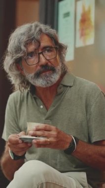 Gri saçlı, sakallı, sıradan kıyafetler giyen düşünceli orta yaşlı bir adam sokak kafesinde oturuyor. Gözlüklü olgun beyefendi aromalı kahve içer.