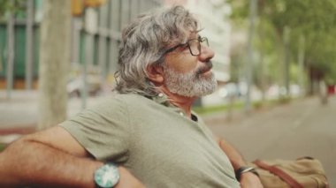 Gri saçlı, sakallı, sıradan kıyafetler giyen orta yaşlı bir adam bankta oturuyor. Gözlüklü olgun beyefendi bankta dinleniyor.