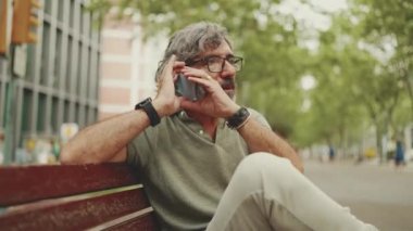 Friendly middle-aged man with gray hair and beard wearing casual clothes sits on bench and uses mobile phone. Mature gentleman in eyeglasses talking on cellphone