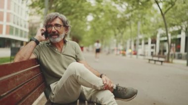 Dost canlısı, orta yaşlı, gri saçlı ve sakallı sıradan kıyafetler giyen bir adam bankta oturuyor ve cep telefonu kullanıyor. Gözlüklü bir beyefendi cep telefonuyla konuşuyor.