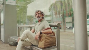 Gri saçlı, sakallı, sıradan kıyafetler giyen düşünceli orta yaşlı bir adam toplu taşıma durağında oturuyor. Gözlüklü ve çantalı bir beyefendi tramvay istasyonunda bekliyor.