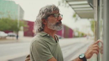 Profile of middle-aged man with gray hair and beard, runs his finger along the city transport schedule. Mature gentleman in eyeglasses with bag on his shoulder is waiting for tram