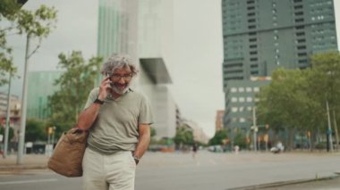 Dost canlısı, orta yaşlı, gri saçlı, sakallı, sıradan kıyafetler giyen ve cep telefonunu kullanan bir adam. Gözlüklü, dışarıda cep telefonuyla konuşan olgun bir beyefendi.