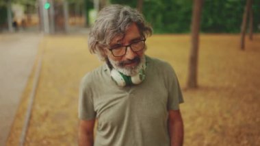 Thoughtful middle-aged man with gray hair and beard wearing casual clothes with headphones around his neck is walking down the street
