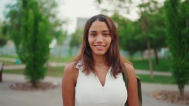 Kahverengi uzun saçlı, bronz tenli beyaz bluzlu güzel kız kameraya bakıyor. Gülümseyen genç bir kadın. Kamera ileri gidiyor.