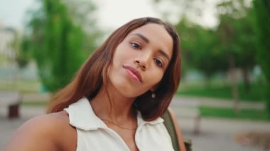 Elbiseli, kahverengi uzun saçlı, bronz tenli, beyaz bluzlu güzel kız cep telefonu kullanıyor. Gülümseyen genç kadın cep telefonundan selfie çekiyor.