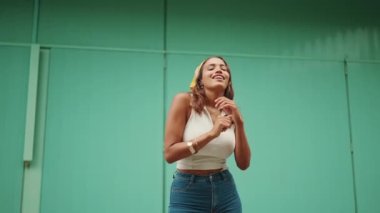Cute tanned woman with long brown hair wearing white top and yellow bandana listens to music in wireless headphones and dances on blue wall background. Camera moves around