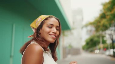 Uzun kahverengi saçlı, beyaz tişörtlü, sarı bandanalı hoş bir bayanın yakın çekimi kablosuz kulaklıkla müzik dinliyor ve mavi duvar arkasında dans ediyor. Yan hareket