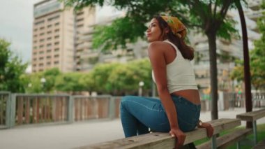 Gülümseyen, bronz tenli, uzun kahverengi saçlı, beyaz bluzlu, sarı bandanalı, kentin arka planında oturan hoş bir kadın profili.