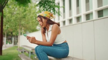 Uzun kahverengi saçlı, beyaz bluzlu ve sarı bandanalı hoş bir kadının profili şehir planındaki bankta cep telefonuyla oturuyor. Kamera ileri gidiyor.