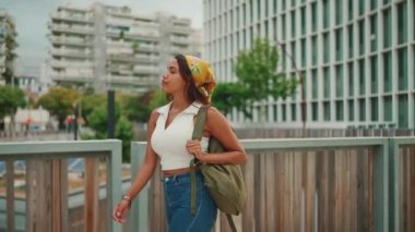 Profile of cute tanned woman with long brown hair wearing white top and yellow bandana walks on bridge with backpack on her shoulder and looks at modern city