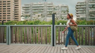 Profile of cute tanned woman with long brown hair wearing white top and yellow bandana walks on bridge with backpack on her shoulder and looks at modern city