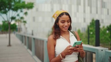 Cute tanned woman with long brown hair wearing white top and yellow bandana walks on bridge with backpack on her shoulder and cell phone in her hand, looks at modern city