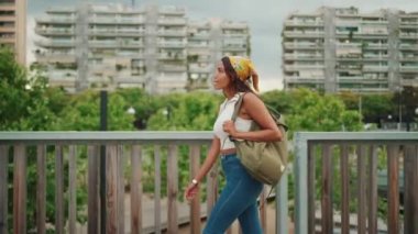 Profile of cute tanned woman with long brown hair wearing white top and yellow bandana walks on bridge with backpack on her shoulder and looks at modern city
