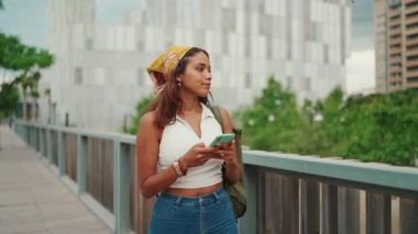 Cute tanned woman with long brown hair wearing white top and yellow bandana walks on bridge with backpack on her shoulder and cell phone in her hand, looks at modern city