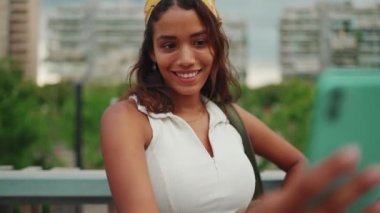 Uzun kahverengi saçlı, beyaz bluzlu ve sarı bandanalı hoş bir kadın modern şehir arka planında köprü üzerinde dururken selfie çekiyor.