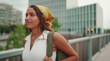 Clouse-up, cute tanned woman with long brown hair wearing white top and yellow bandana walks on bridge with backpack on her shoulder and looks at modern city