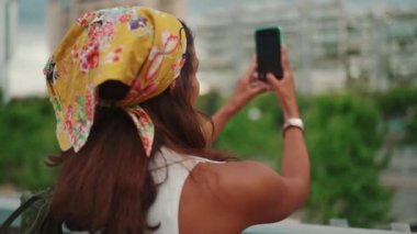 Clouse-up, tanned woman with long brown hair wearing white top and yellow bandana standing on bridge taking photo of modern city