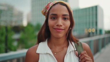 Clouse-up, cute tanned woman with long brown hair wearing white top and yellow bandana posing while standing on bridge. Smiling girl looking at the camera