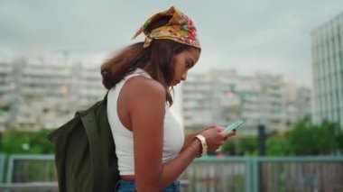 Cute tanned woman with long brown hair wearing white top and yellow bandana with backpack on her shoulder is using mobile phone on modern building background. Moving around