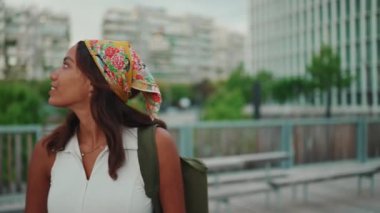 Clouse-up, cute tanned woman with long brown hair in white top and yellow bandana with backpack on her shoulder and looks around the modern city. Camera moving around