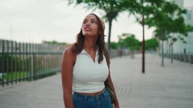 Cute tanned woman with long brown hair in white top and yellow bandana with backpack on her shoulder and looks around the modern city. Camera moving around