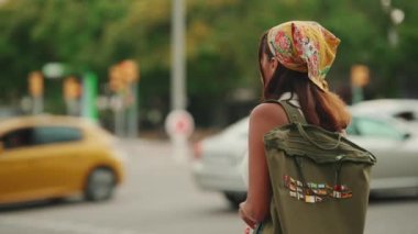 Cute tanned woman with long brown hair in white top and yellow bandana with backpack on her shoulders stands at crosswalk on the road background. Back view. Camera moving around